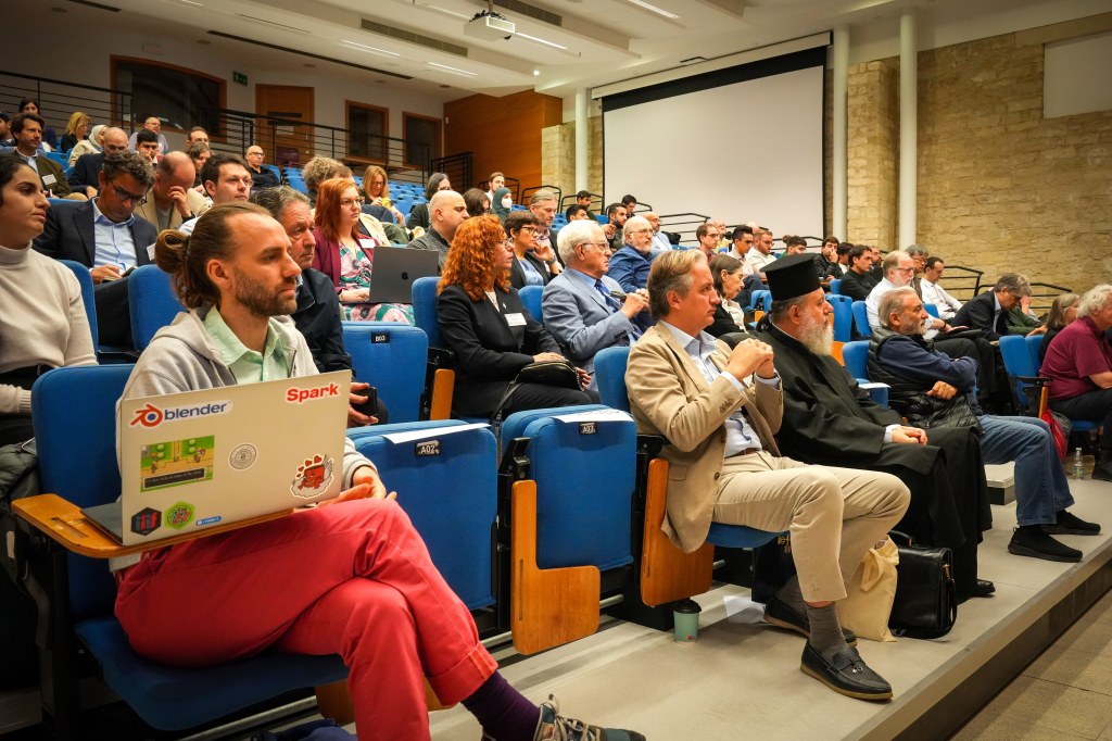 A conference audience seated in a lecture hall, featuring individuals engaged in discussions and looking towards a presenter, with a diverse mix of attendees including men and women.