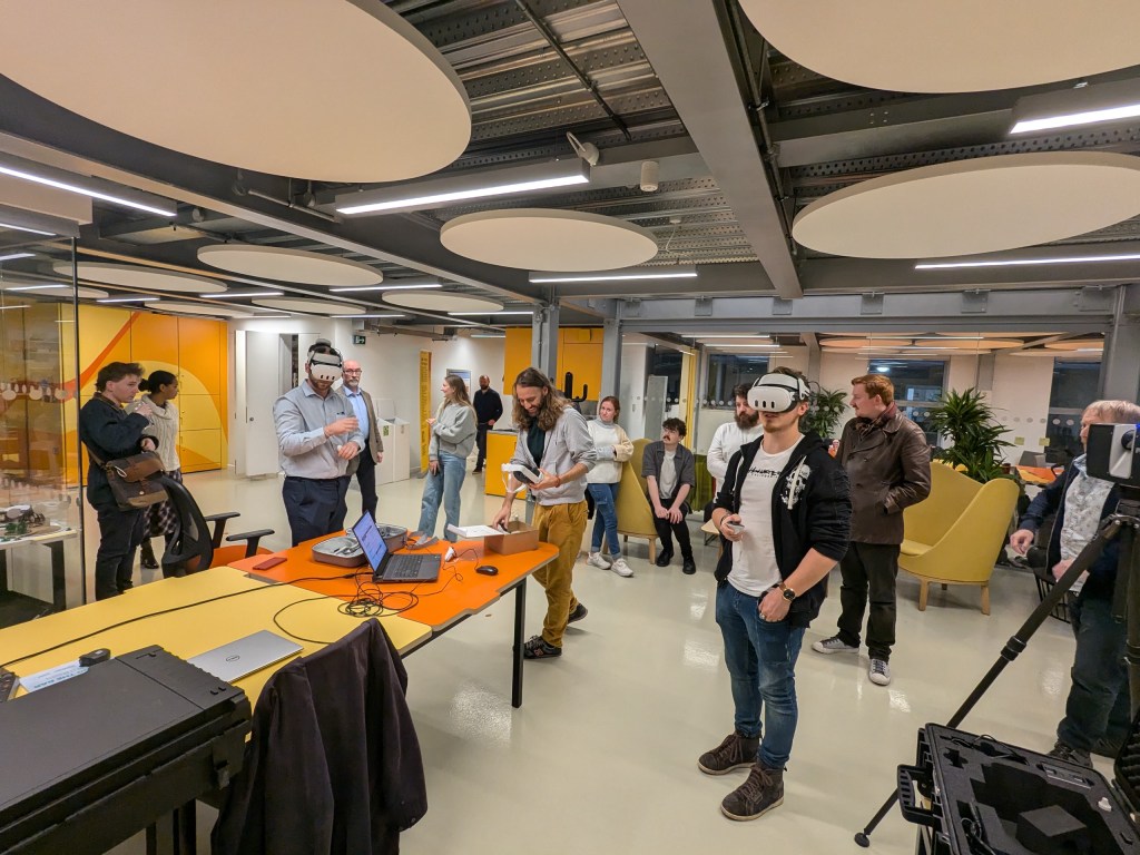 A group of individuals participating in a workshop, with some wearing virtual reality headsets. They are gathered around a table with a laptop and various equipment in a modern, brightly lit environment.