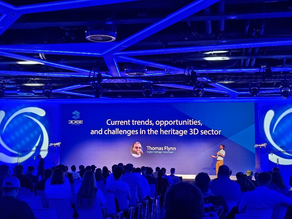 A presentation at a conference featuring Thomas Flynn discussing trends, opportunities, and challenges in the heritage 3D sector, with an audience engaged in the foreground.